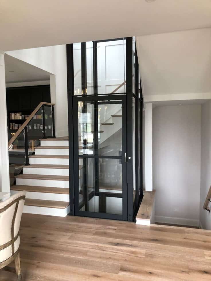 Hình ảnh thang máy mini lắp đặt cạnh cầu thang trong nhà phố Quận Thủ Đức. Kiểu dáng hiện đại với khung thép đen sang trọng, tạo nên sự hài hòa với sàn gỗ tự nhiên và nội thất ấm cúng.