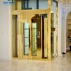 Thang máy cao cấp với lớp phủ inox vàng gương, nổi bật trong không gian biệt thự trần cao, sàn đá tự nhiên.