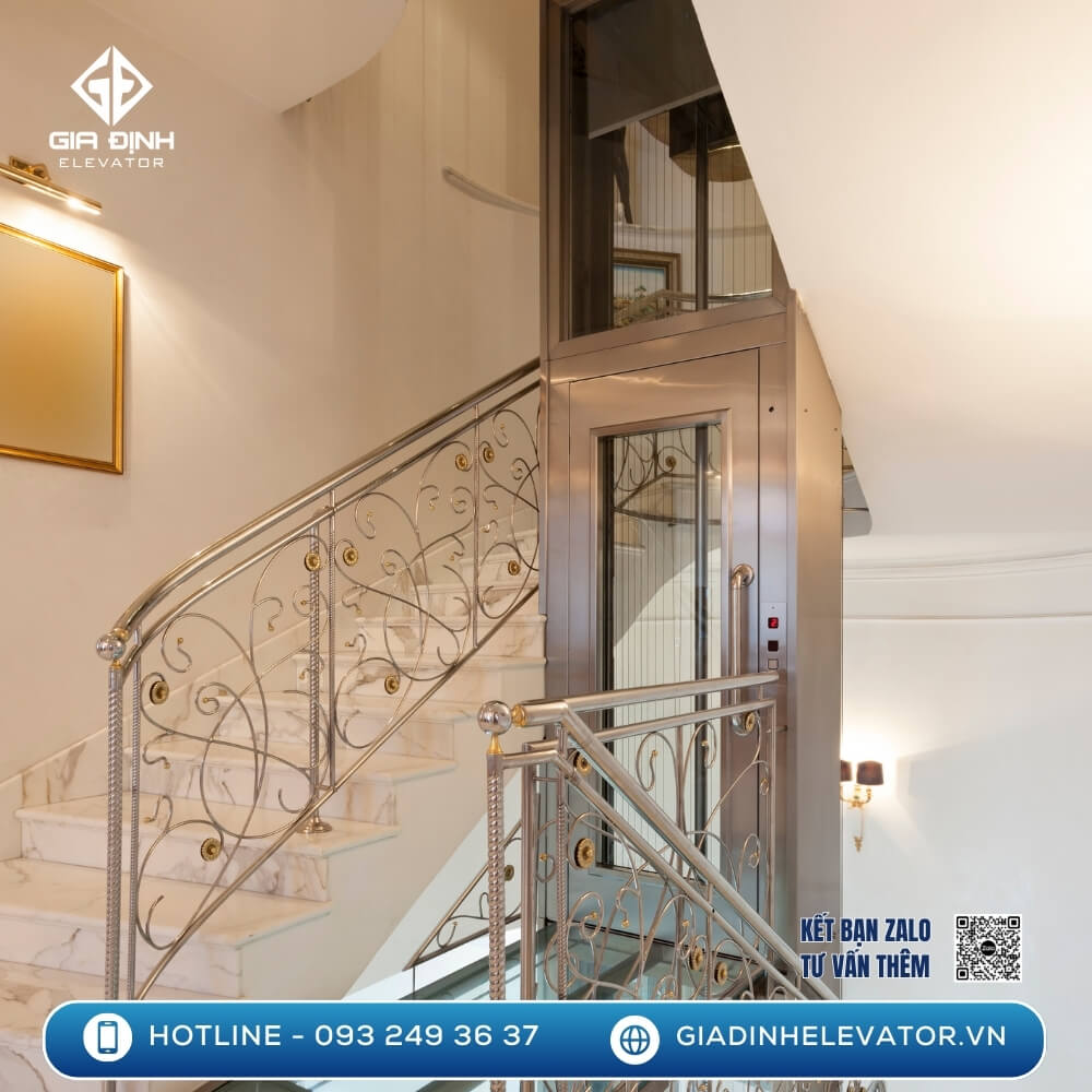 Mẫu thang máy mini đặt giữa lối đi cầu thang nhà ống, với lan can sắt nghệ thuật và không gian nội thất cao cấp.